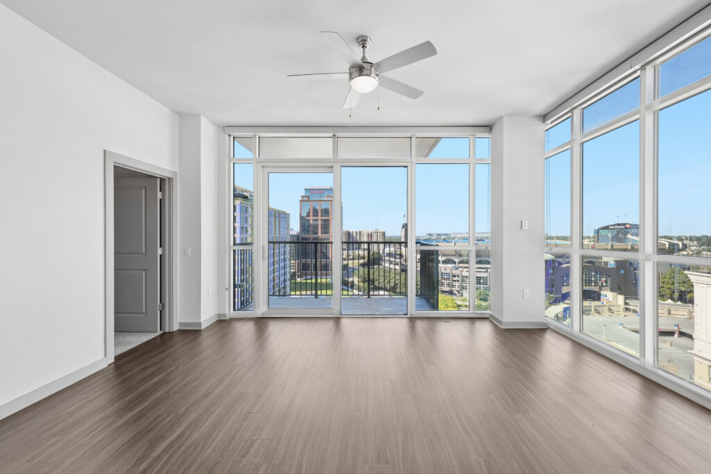 Modern empty apartment room with large windows, wood flooring, a ceiling fan, and a balcony overlooking a cityscape with buildings and a distant river or bridge.