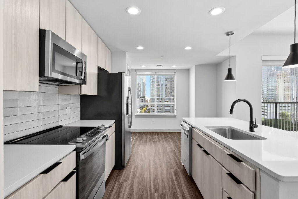 Modern kitchen with light wood cabinets, stainless steel appliances, white countertops, subway tile backsplash, pendant lights, and a large window offering a city view.