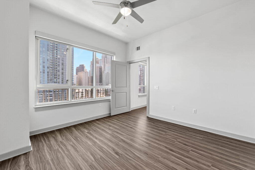 Empty room with wood flooring, white walls, a ceiling fan, large window showing a city skyline, and an open door. Bright natural light fills the space.