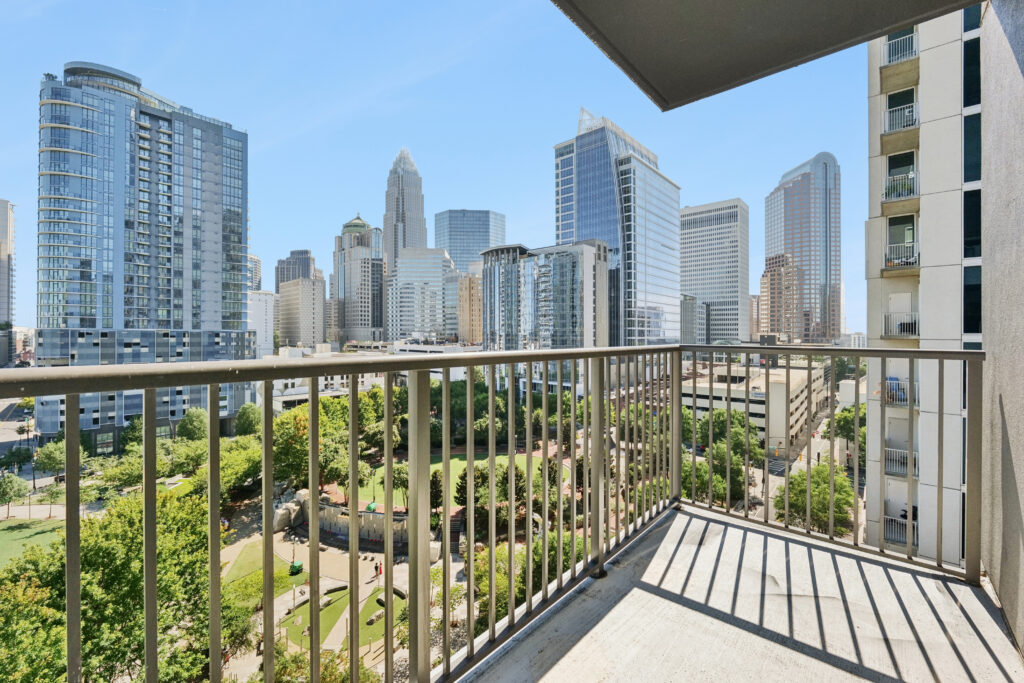A sunny city view from a high-rise balcony, showing modern skyscrapers, green park areas below, and blue sky in the background.