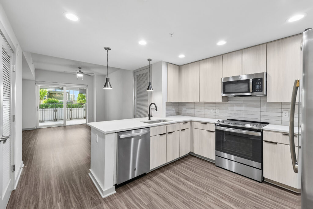 Modern kitchen with light wood cabinets, stainless steel appliances, pendant lighting, and a view into a bright living area with large windows and a balcony.