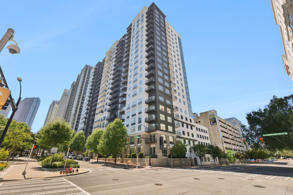 A modern high-rise apartment building with balconies on a sunny day, located at a city street corner with trees, crosswalks, and nearby buildings visible.