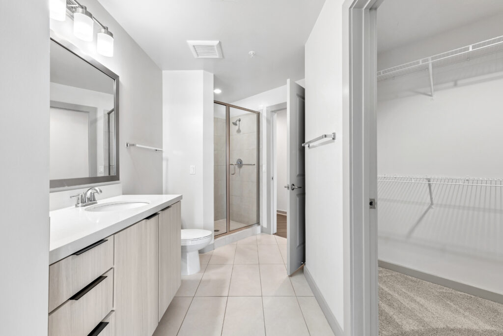 Modern bathroom with a double sink vanity, toilet, glass shower, and adjacent walk-in closet featuring wire shelving. The space has neutral colors and bright lighting.