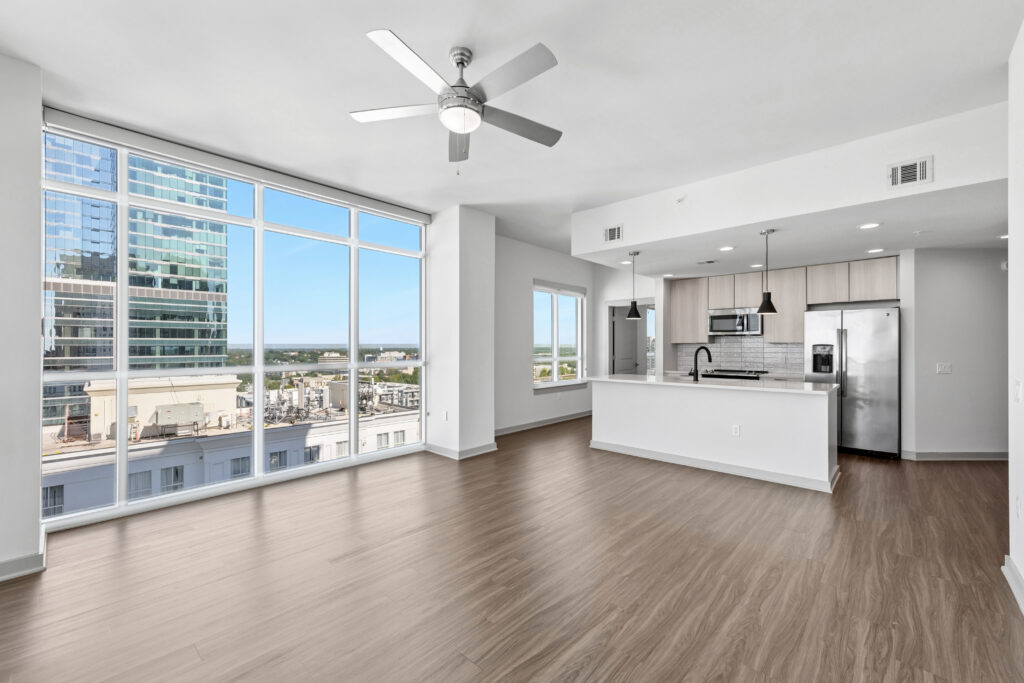 Modern, unfurnished apartment with large windows, wood floors, a ceiling fan, and an open kitchen featuring stainless steel appliances and light cabinets. Cityscape and blue sky visible through the windows.