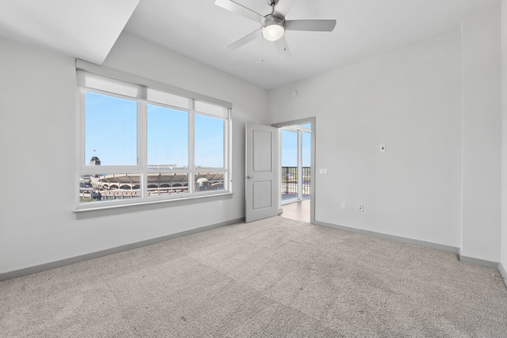 Bright, empty bedroom with beige carpet, large windows showing a city view, white walls, a ceiling fan with light, and an open door leading to a balcony.