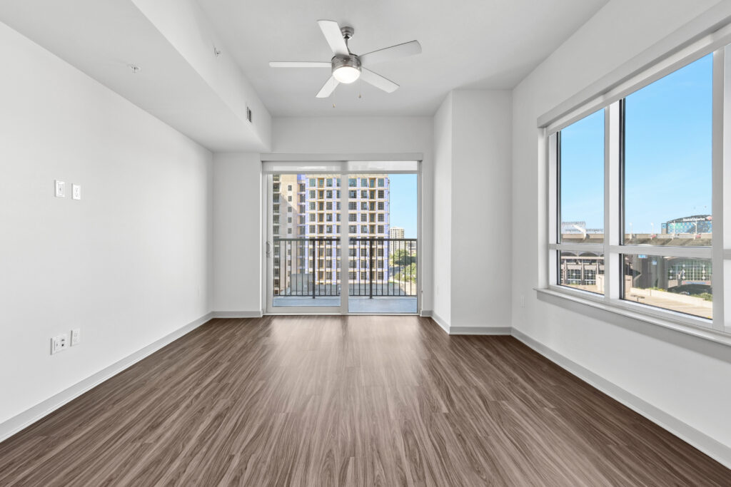 Modern empty apartment room with wood floors, white walls, ceiling fan, large windows, and a sliding glass door leading to a balcony with a cityscape view.