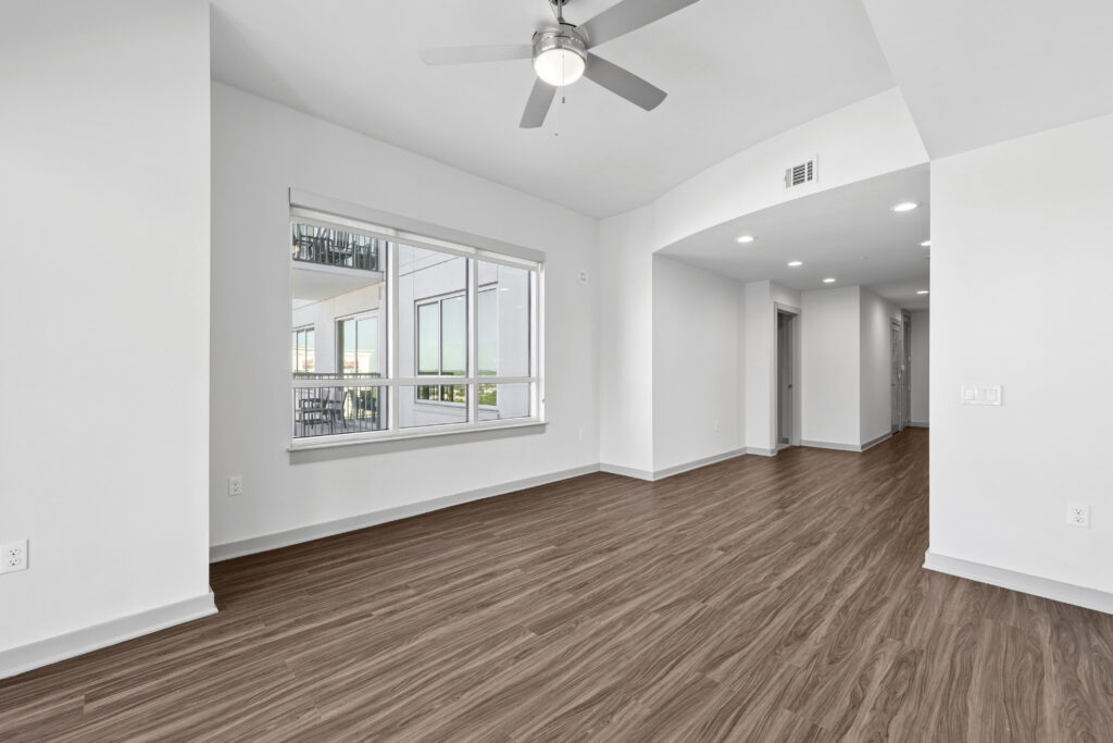 A modern, unfurnished apartment room with wood floors, white walls, ceiling fan, large window, recessed lighting, and a hallway leading to other rooms.