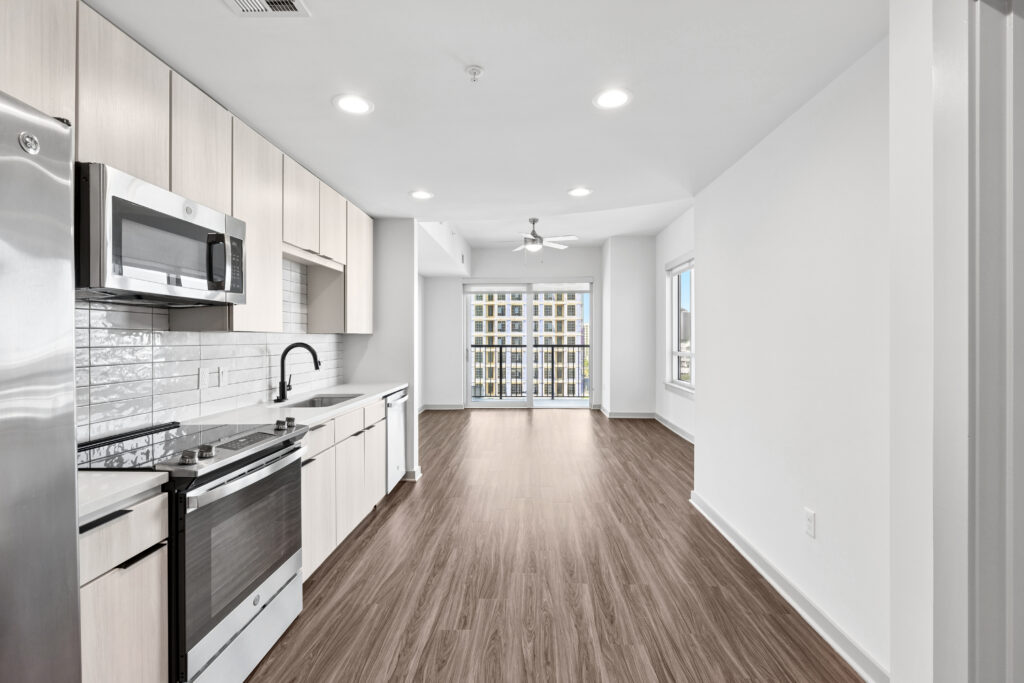 Modern kitchen with light wood cabinets, stainless steel appliances, and white subway tile backsplash, opening to a bright living area with wood floors, ceiling fan, and large windows.
