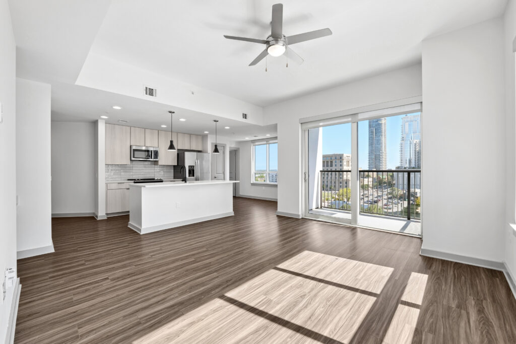 Modern apartment with wood floors, open kitchen with white cabinets and stainless steel appliances, ceiling fan, and large windows leading to a balcony with a city view.