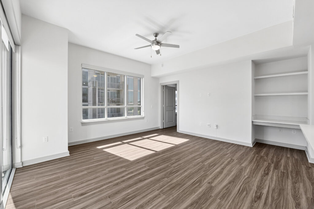 Bright empty room with large window, ceiling fan, wood-style flooring, built-in shelves, and desk area. White walls and plenty of natural light create a spacious feel.